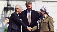 A stolid balding man in a dark suit on the left shakes the hand of a smiling man in traditional Arab headdress on the right. A taller, younger man stands with open arms in the center behind them.