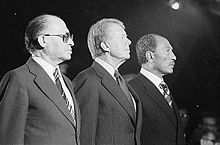 Three middle-aged, suited men standing in the spotlight, seen from the side. The men are looking away from the camera, with serious expressions. In the background are several other people, out of focus.